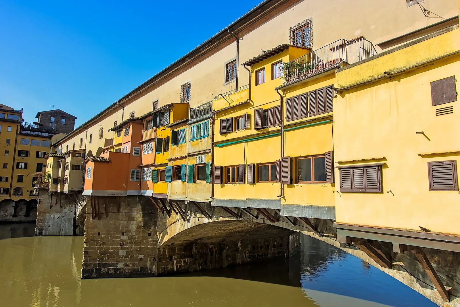 Il Ponte Vecchio di Firenze visto dal basso con le botteghe colorate gialle e arancioni sospese sull'Arno e il corridoio superiore, sotto un cielo azzurro