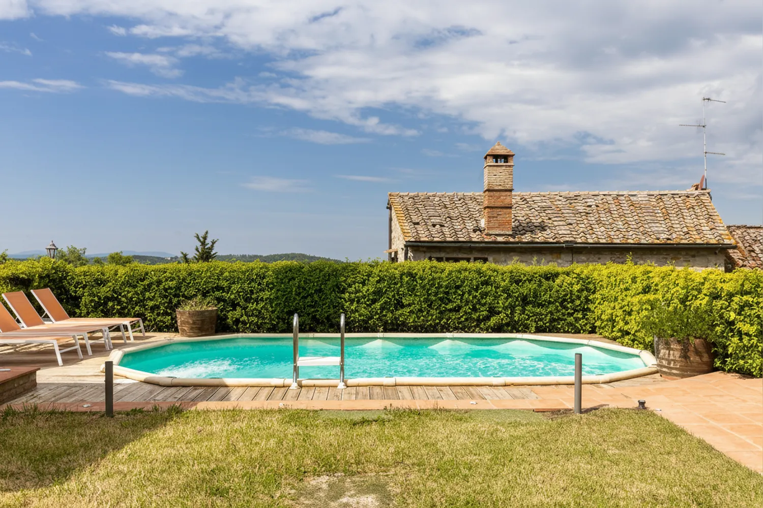 La piscina con le sdraio arancioni, la siepe e il tetto di un casale vicino